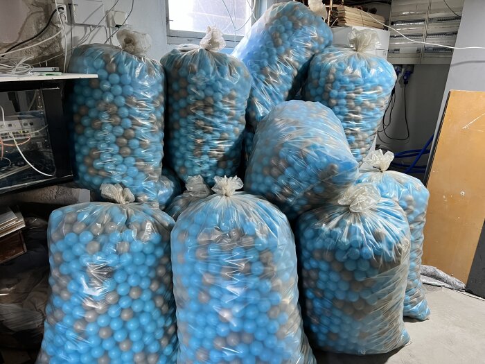 19 plastpåsar fyllda med blå och grå bollar staplade i ett rum med elektronik och kablar på väggen.