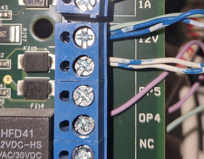 En närbild av en kopplingsplint med blå och vita inplastade kablar anslutna, märkta med 12V, OP5 och OP4.