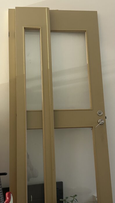 Gulmålad dörr med glasfönster och metallhandtag, uppställd mot en vägg.