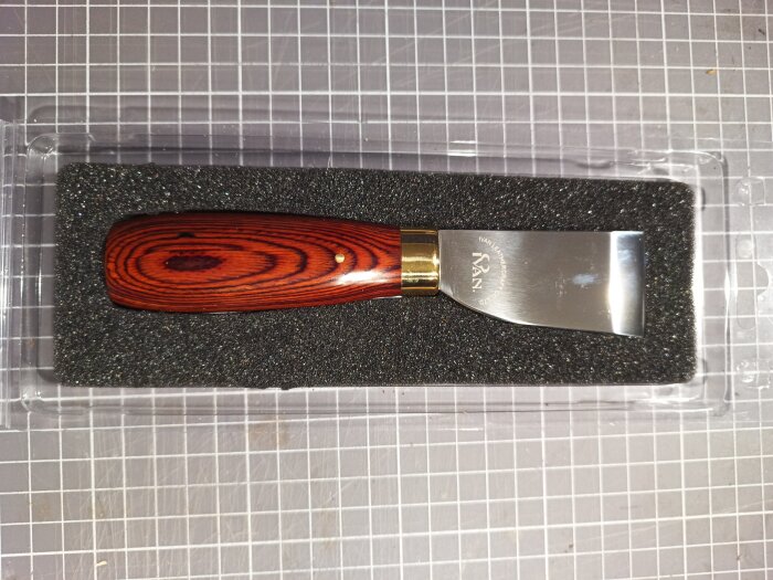 Kniv för att tunna ut läder med trähandtag och skarpt blad, ligger i svart skumfodral på ett rutigt underlag.