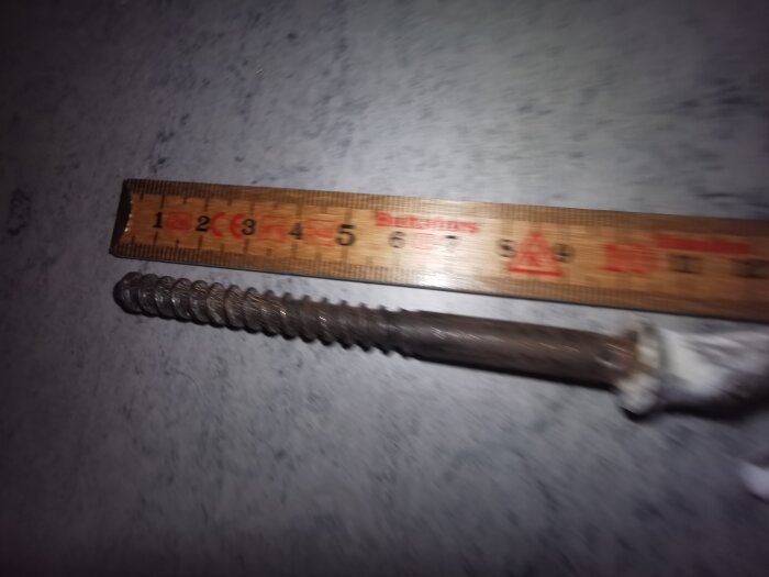 Stor metallskruv bredvid en trälinjal med markeringar. Skruven har grova gängor och ser ut att vara cirka 9 cm lång.