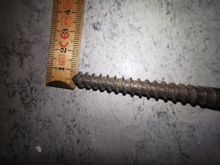 Rostig skruv bredvid en linjal på ett gråprickigt underlag, försök att identifiera skruvtjocklek och gänga i äldre konstruktion.