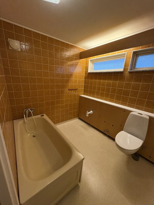 Badrum med orange kakelväggar, beige badkar, toalett och skåp. Smala fönster ovanför skåpen, golv i ljus nyans.