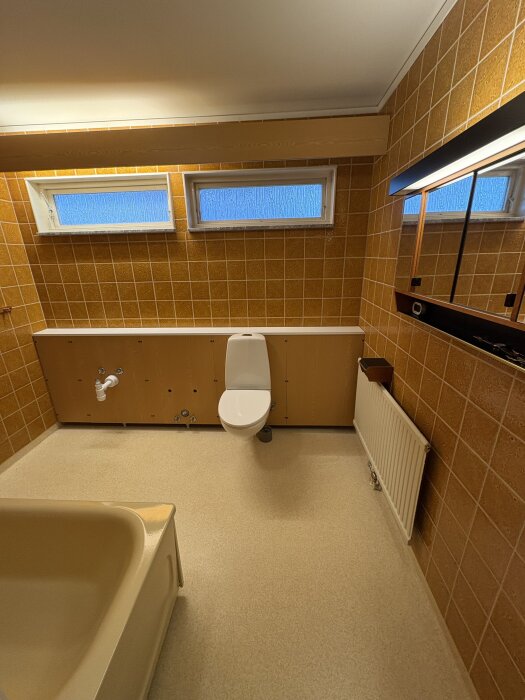 Badrum med orange kakel, toalettstol, badkar och två små fönster. Spolrör och rör för tvättställ synliga. Spegel ovanför radiator.