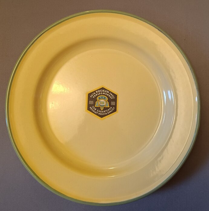 Gul emaljtallrik från Kockums med märkning i mitten, tillverkad i Ronneby före 1971.