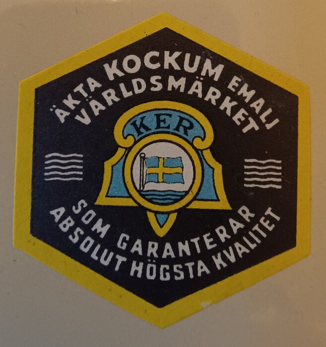Märke för Kockums emalj, med text "Äkta Kockum emalj världsmärket" och "Som garanterar absolut högsta kvalitet", inklusive en symbol med en flagga.