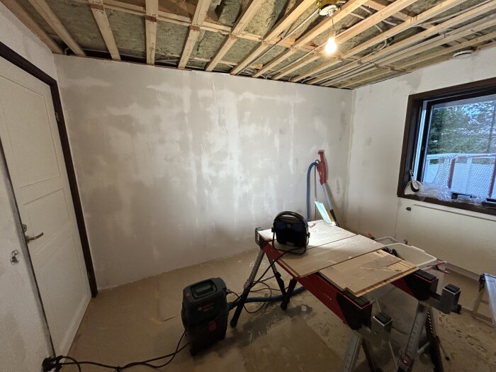 Rum under renovering med målarbord och verktyg, isolering i taket och en bygglampa.
