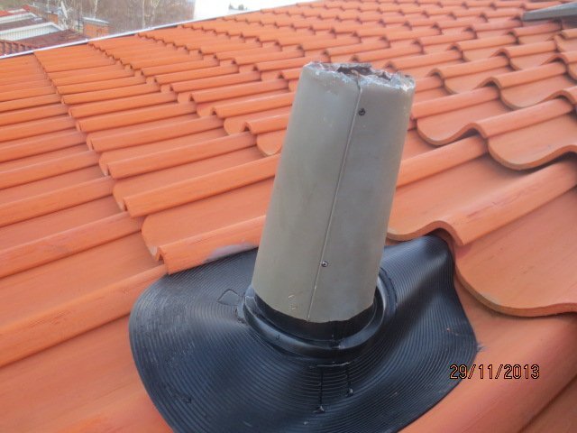 En avluftningsventil genom yttertak med röd takpanna och en plastkrage runt röret, fotograferad på nära håll.