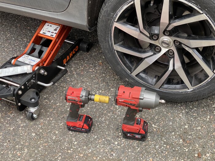 Bilverkstadsscen med en domkraft under bilen och två röda elektriska mutterdragare framför ett bildäck.