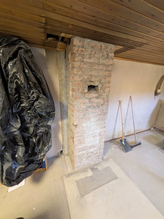 Murad skorsten i ett rum med träpanel i taket, omgiven av verktyg och svart plastskydd.