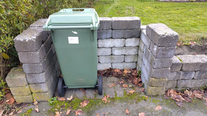 Grönt sopkärl placerat framför en staplad stenmur som skapar ett utrymme vid tomtgränsen. Lövgolv och mossa runt omkring.