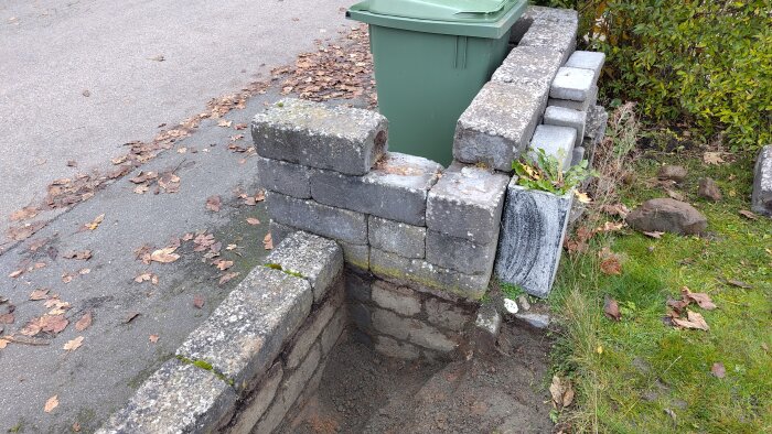 Inbyggd stenmurskonstruktion vid tomtgräns med bredvidstående sopkärl. Lövprydd asfaltsyta och gräsmatta omger muren.