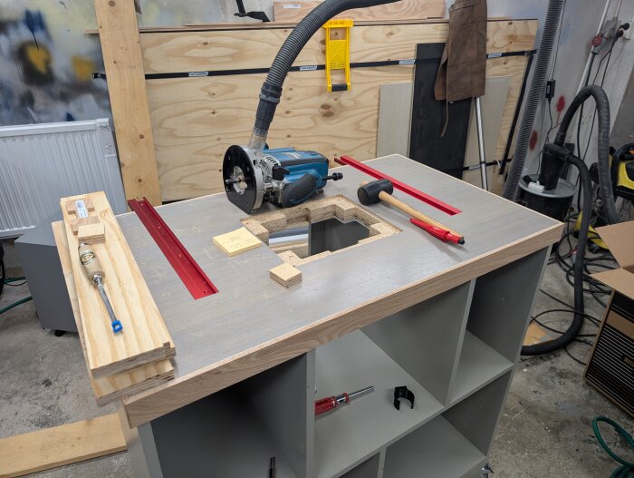 Fräsbord med infälld fräs och röda skenor; verktyg som gummiklubba, skruvmejsel och en slanghållare i taket syns i verkstaden.