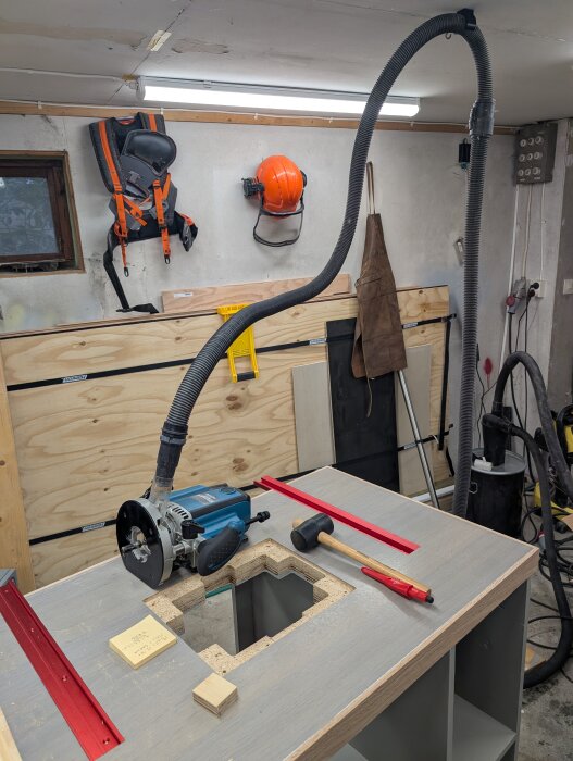 Fräsbord med monterad slanghållare i taket, rödfräst spår för anhåll, verktyg på bordet i verkstadsmiljö.