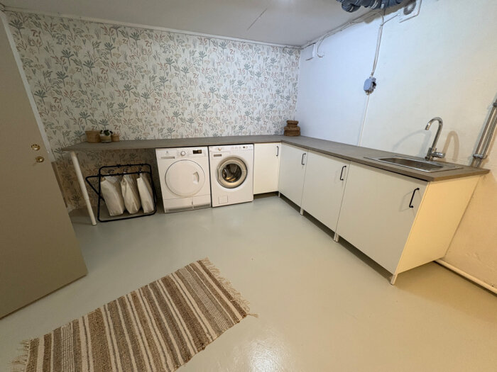 Tvättstuga med tvättmaskin, torktumlare, bänk med diskho, korgar och förvaringsskåp. Ljus tapet i bakgrunden och en randig matta på golvet.