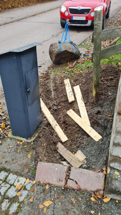 En stor sten har flyttats med en bil och blå remmar, omges av jord och träplankor, förbereds för sopsorteringstunnor.