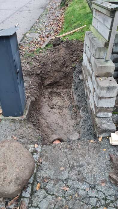 En nygrävd grop vid sidan av en gångväg med stenar och en stapel av byggblock. En spade ligger på gräsmattan bredvid.