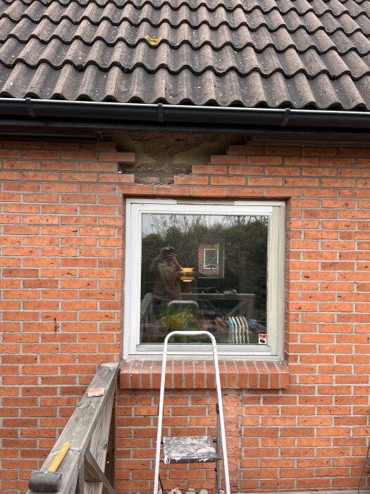 Fasad av rött tegel med ett fyrkantigt vitt fönster, ovanför syns borttaget tegel för att exponera gammal tegelbalk. En stege står mot väggen.