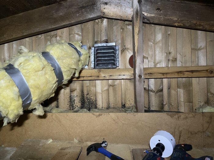 Gullfiberisolerat rör i en träkonstruktion på vind, med tejp och verktyg bredvid.