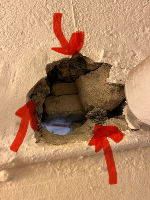 Hål i väggen markerat med röda pilar, visar exponerat murverk under vit yta, eventuellt för reparation eller inspektion.