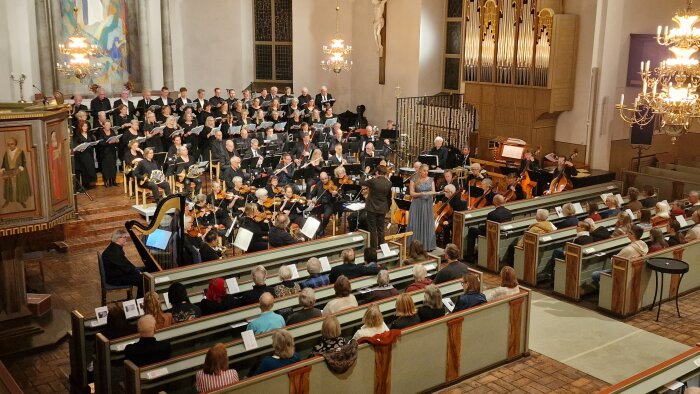 Kör och orkester uppträder i kyrka inför publik, dirigent leder framträdandet.