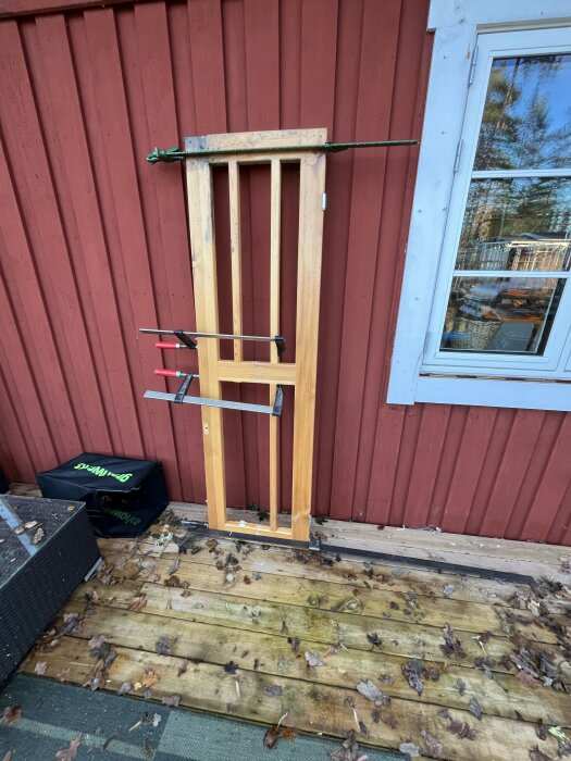 Trädörrstomme med spännband och klämmor, stående mot en röd träfasad, omgiven av löv på altanen.