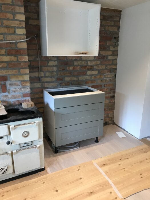 Ett hörn av ett kök med en gammal vedspis, en bröstpanel av tegel, ett grått bänkskåp och ett tomt väggskåp över skivan.