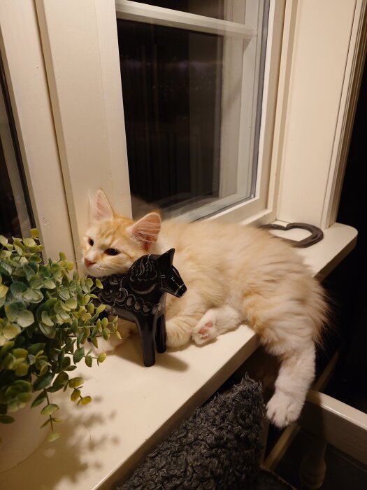 Ljus kattunge vilar på en fönsterbräda bredvid en svart dalahäst och en grön växt.