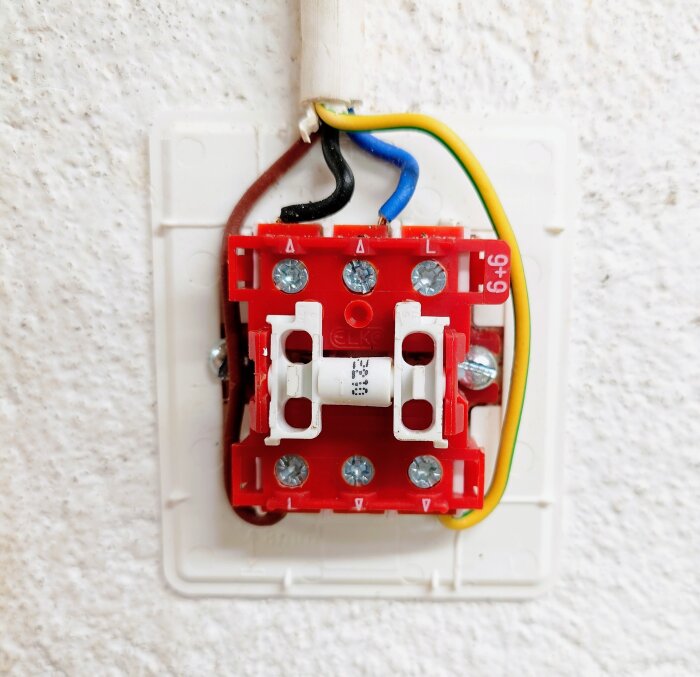 Elkopplingsdosa med flera kablar synliga: svart, blå, gul/grön, anslutna till en röd brytare monterad på vägg.