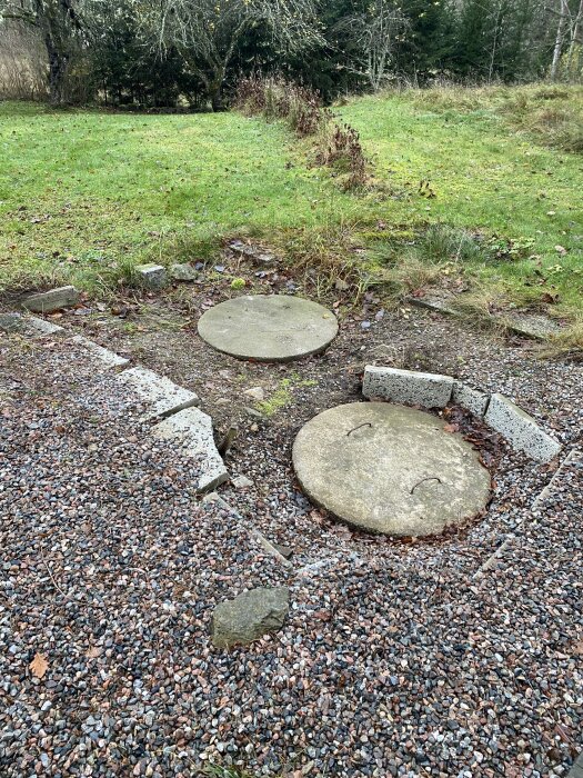 Betonglock för avloppstank placerade i marknivå, omgivna av grus och gräs, intill en grön uteplats med buskar i bakgrunden.