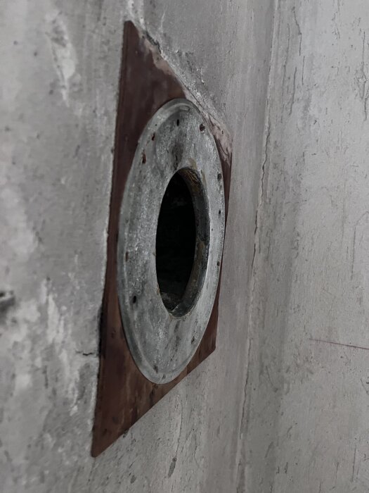 Gammal tallriksventil i en vägg gjord av betong, rödbrun fyrkantig platta runtom, ventilen är rostig och delvis öppen.
