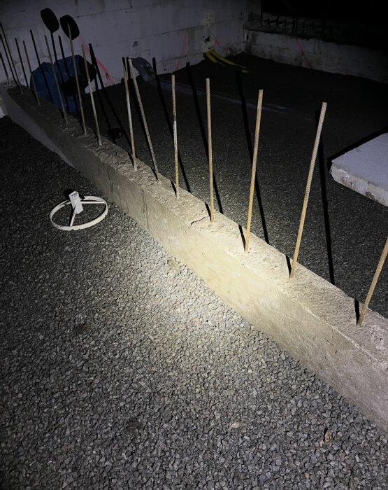 Grundmur i betong med armeringsjärn som sticker upp, placerad på grus. Omgivande utrustning synlig i bakgrunden.