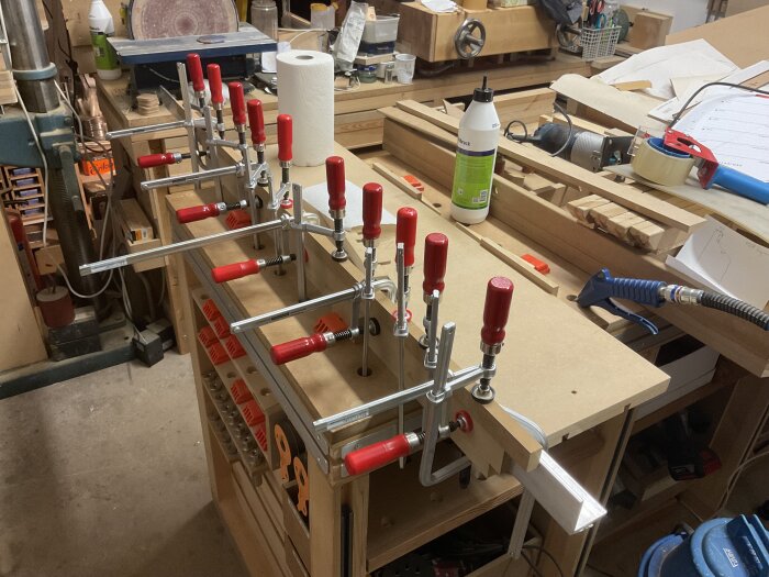 Ett arbetsbord i en verkstad med flera träklampar fastsatta med långa, röda skruvtvingar. Verktyg och material ligger på bordet i bakgrunden.