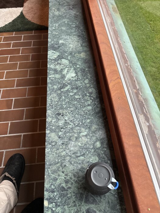 Grön marmorfönsterbräda vid ett fönster, delvis slipad, med en liten svart föremål ovanpå och rödbruna golvplattor bredvid.