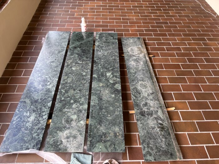 Fyra gröna marmorliknande fönsterbrädor, två slipade och två med synliga ringar, ligger på ett golv av bruna tegelplattor.