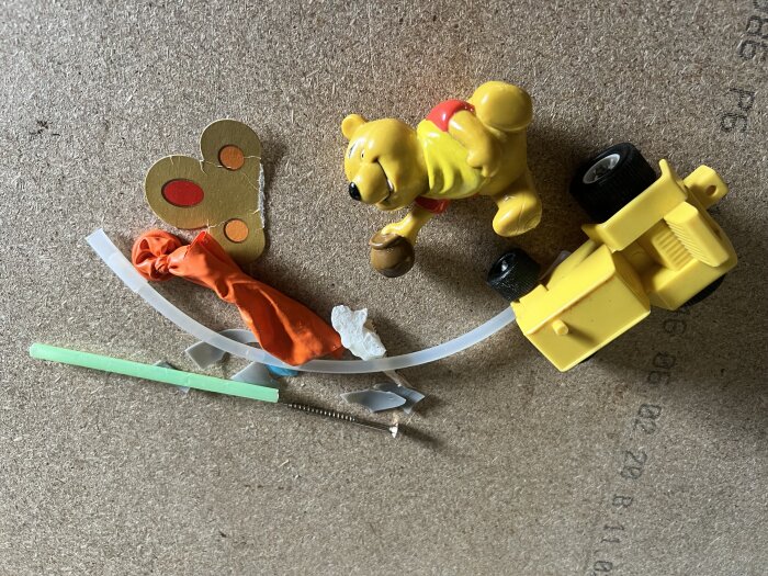 Gula leksaker inklusive Nalle Puh-figur och liten gul traktor på golvet omgiven av blandade småsaker som papper, sugrör och en ballong.