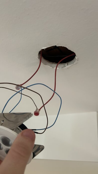 Hand som håller en vit downlightlampa nära tak med synliga blå och röda kablar från ett hål.