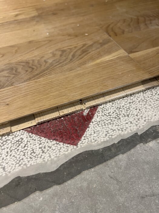 Trägolv ovanpå en underlag bestående av betong och vit cellplast med del av röd markering synlig.