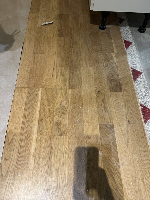 Ljusa trägolv med mönster av stavparkett, delvis skuggat, sett under vit möbel och bredvid röd-vit kakelplatta.