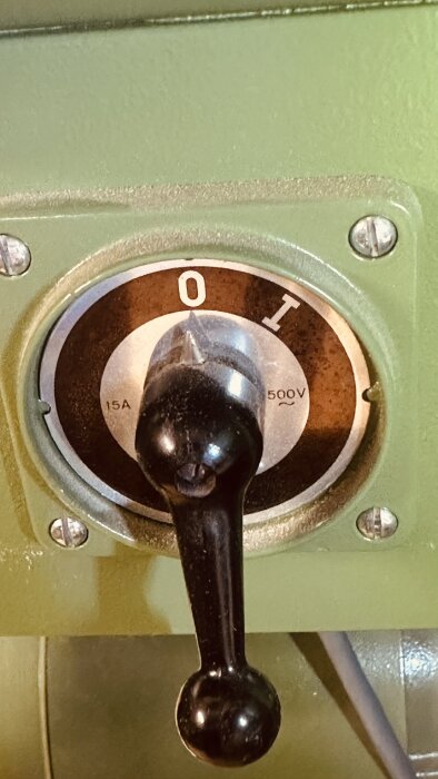 Närbild på en svart strömbrytare inställd på "0" med markeringar för 15A och 500V tydliga, monterad på en grön maskin.