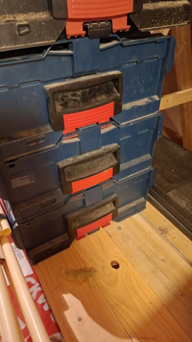 Tre staplade verktygslådor med röda handtag på ett träbord i ett vindsutrymme.