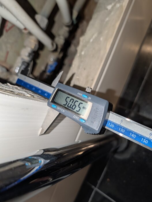 Vägg med digital skjutmått visar tjocklek på 50,65 mm för kakel och trippelgips i badrum med rör och beslag i bakgrunden.