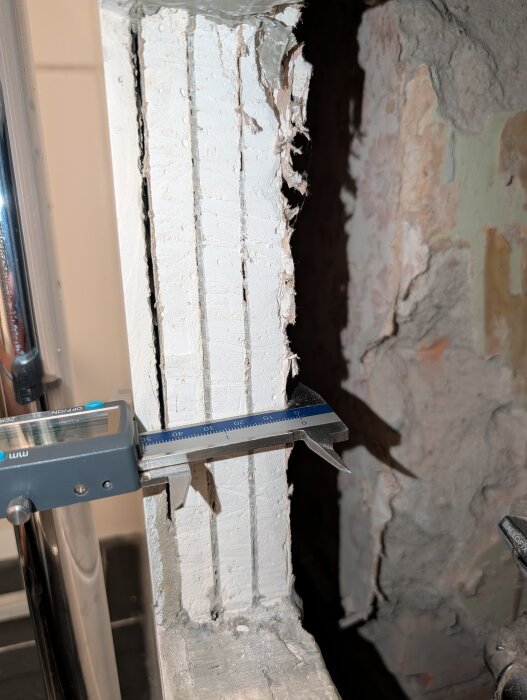 Digital skjutmått mäter gipsskivans tjocklek på en vägg med flera lager, omgiven av trasigt material och kakel.