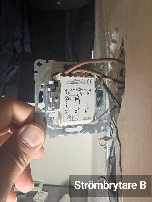 En strömbrytare som hålls i en hand, inbyggd i väggen med synliga kablar i brunt och svart, etiketterad "Strömbrytare B".