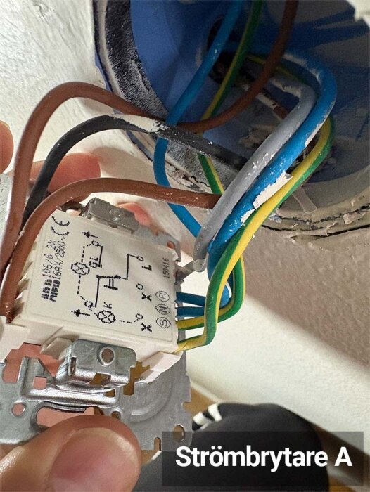 Närbild på strömbrytare A med flera färgade elkablar och ett kopplingsschema synligt. Bakgrunden visar en del av väggen.