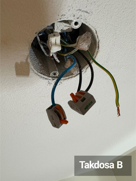 Takdosa B med kablar, där två är kopplade via Wago-kopplingsklämmor, en grön/gul, en svart och en blå kabel är synliga i eldosan.
