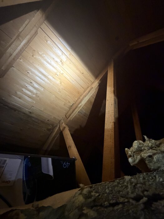 Loft med träbalkar i takkonstruktion och isolering i förgrunden, del av renoveringsprojekt.