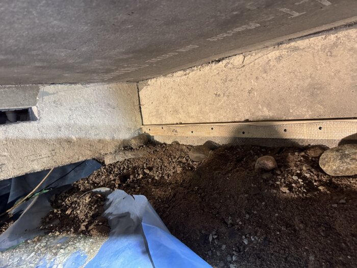 Bild av ett utrymme under hus med installerad järntrådduk fäst vid glespanel; marken täckt med jord och några stenar synliga.