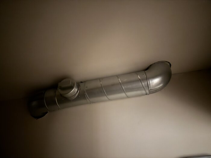 Metallventilationsrör med böjd del monterat i tak, sett i ett svagt upplyst rum.