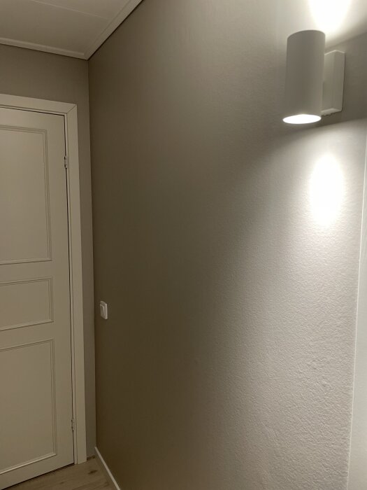 Vägg i hall med dörr, renoverad med spackel och tapet, och en vägglampa lyser upp hörnet.